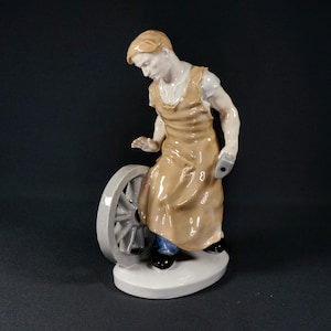 Może przedstawiać: Porcelanowa figurka przedstawiająca osobę w beżowym fartuchu nałożonym na białą koszulę i niebieskie spodnie, stojącą obok szarego koła. Postać trzyma w dłoni małe szare narzędzie i jest umieszczona na białej owalnej podstawie na ciemnym tle.