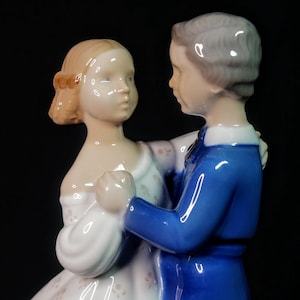 May include: A porcelain figurine depicting a dancing couple. The woman wears a white dress with floral accents, and the man is in a blue jacket. The figurine has a glossy finish and is set against a black background.