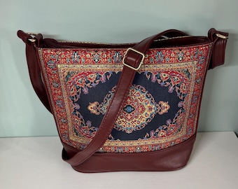 May include: A burgundy shoulder bag with a rectangular, patterned front panel. The panel features a navy blue background with a central medallion and ornate red, yellow, and blue floral designs. The bag has a matching burgundy base and strap.