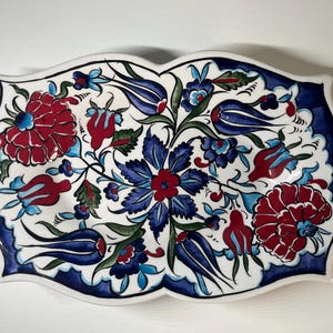 May include: A white ceramic serving dish with a scalloped edge, decorated with a vibrant floral pattern. The design features red and blue flowers with green leaves, set against a white background, and outlined with a dark blue border.