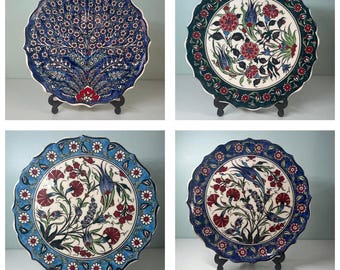 May include: Four decorative ceramic plates with intricate floral designs. Each plate features a unique color scheme: blue, green, and white, with red and blue floral patterns. The plates are displayed on black stands.