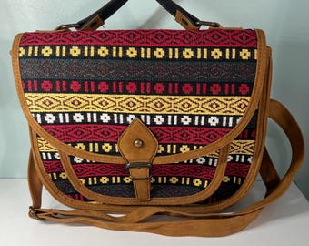 May include: A shoulder bag with a woven fabric flap in red, black, and yellow geometric patterns. The bag has a brown leather trim, a brown leather strap, and a black handle. The bag is closed with a brown leather buckle.