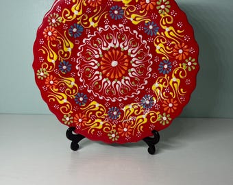 May include: A vibrant red decorative plate with a scalloped edge. The plate features a colorful floral design in yellow, orange, blue, and white. The plate is displayed on a black stand, showcasing its intricate patterns and bright colors.