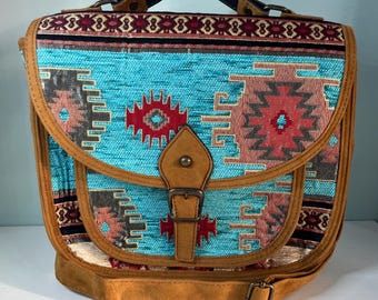 May include: A turquoise and brown satchel bag with a Southwestern pattern. The bag features a flap closure with a buckle, a top handle, and a shoulder strap. The bag's trim is brown, and the handle is black.