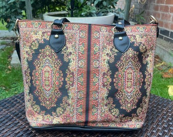 May include: A rectangular tote bag with a patterned design. The bag features a black base with a colorful floral pattern in red, gold, and green. It has black handles and a shoulder strap. The bag is sitting on a brown wicker table.