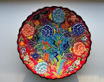 May include: A decorative ceramic plate with a vibrant floral design. The plate features a red background with a scalloped edge and is adorned with colorful flowers in shades of blue, orange, and yellow. The plate measures approximately 12 inches in diameter.