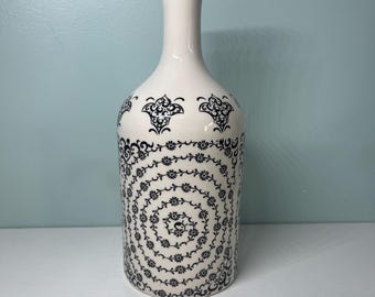 May include: A white ceramic bottle with a cork stopper. The bottle is decorated with black floral patterns, including a spiral design on the body and ornate shapes near the top. The bottle is on a white surface against a light blue background.