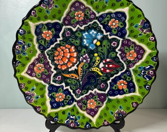 May include: A decorative ceramic plate with a scalloped edge, featuring a vibrant floral design. The plate showcases a green background with orange, blue, purple, and white floral patterns. The rim is black, and the plate is displayed on a black stand.