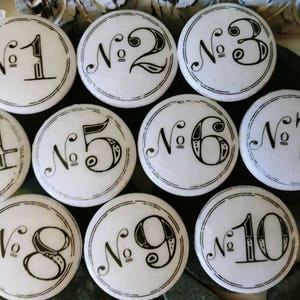 Könnte beinhalten: Zehn weiße runde Keramikknöpfe mit schwarzen Zahlen von eins bis zehn. Jeder Knopf hat einen schwarzen Rand und ein dekoratives Design um die Zahl.