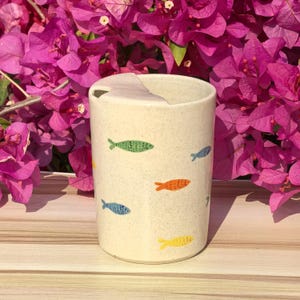 Puede incluir: Un vaso de cerámica beige con tapa, decorado con ilustraciones de peces de colores. Los peces son en tonos verdes, naranjas, azules y amarillos. El vaso es cilíndrico y parece hecho a mano.