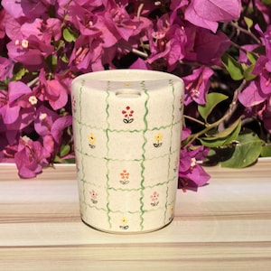 May include: A cylindrical ceramic container with a beige background, decorated with a green grid pattern and colourful floral designs. The flowers are red, yellow, orange, and pink. The container has a small hole on the top.