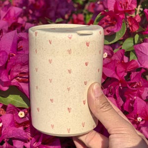 Peut inclure: Un récipient cylindrique en céramique avec une finition beige mouchetée, orné de petits motifs de cœurs roses espacés. Le récipient a un dessus incurvé avec une petite ouverture. Le fond présente des fleurs roses vibrantes.