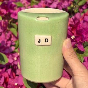 May include: A light green ceramic vase with a beige rim and a small rectangular tag with the letters "J D". The vase is cylindrical with a small opening at the top. The background features vibrant pink flowers.