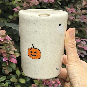May include: A cream-colored ceramic travel mug with a speckled finish, featuring a hand-painted orange jack-o'-lantern, a small white ghost, and the word "boo" written in black ink near the base, with a circular opening on the lid.