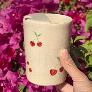 May include: A beige ceramic travel mug with a matching lid. The mug is decorated with hand-painted red cherries and green leaves. The mug is cylindrical and has a capacity of approximately 12 ounces.