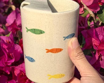Peut inclure: Une tasse en céramique beige avec un couvercle et une fente pour une paille. La tasse est décorée d'illustrations de poissons colorés en vert, bleu, orange et jaune. La tasse est tenue devant un fond de fleurs roses.