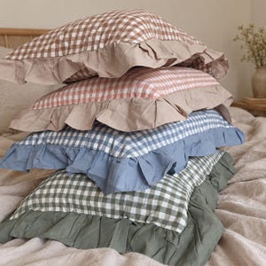 Pode incluir: Uma pilha de quatro almofadas retangulares, cada uma com um padrão de xadrez vichy em castanho, rosa, azul e verde. Cada almofada é rematada com um rebordo de folhos em tecido de cor sólida que combina com o tom principal do vichy.