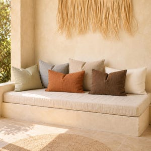 May include: A collection of six rectangular and square throw pillows in various solid colors including sage green, slate gray, burnt orange, beige, dark brown, and off-white, arranged on a long, low, built-in bench with a matching light beige cushion.