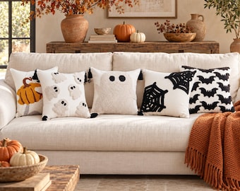 May include: A set of five square throw pillows arranged on a beige sofa. The pillows feature tufted designs including a pumpkin, multiple small ghosts, a large central ghost, black spiderwebs, and a repeating pattern of black bats on white fabric.