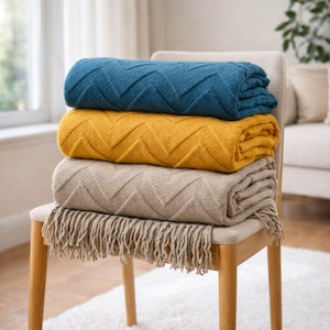 Op de afbeelding: Een stapel van drie opgevouwen dekens in teal, mosterdgeel en beige, elk met een zigzagpatroon. De onderste deken heeft franjes. De dekens liggen op een houten stoel met een neutraal gekleurde zitting.