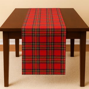 May include: A red tartan table runner with a traditional plaid design. The runner has a vibrant red base, overlaid with lines in yellow, green, and navy blue. It is placed on a dark brown wooden table.