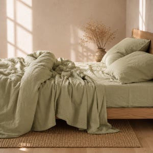 May include: A bed with sage green bedding, including a comforter, fitted sheet, and two pillows. The bed is made of light-colored wood and sits on a woven rug. A vase with dried flowers is on the nightstand.