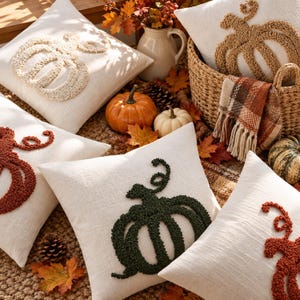 Könnte beinhalten: Eine Sammlung quadratischer, cremefarbener Dekokissen mit texturierten Kürbismotiven in Creme, Dunkelgrün und Rostorange. Die Kissen sind zusammen mit dekorativen Kürbissen, Herbstlaub und einem geflochtenen Korb arrangiert.