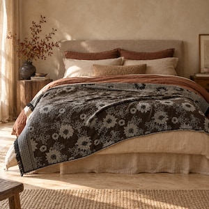 Könnte beinhalten: Eine dunkel anthrazitfarbene Tagesdecke mit wiederkehrendem weißem Blumenmuster und Fransen an den Rändern, drapiert über einem Bett mit beigen und rostfarbenen Laken, inklusive mehrerer Kissen und einem strukturierten Nackenkissen.