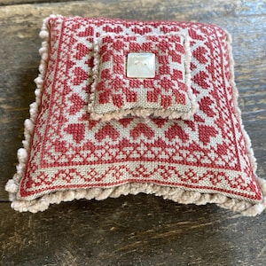 May include: A small, square pincushion with a red and beige cross-stitch pattern. The design features hearts and geometric shapes. A smaller, matching cushion is attached on top, secured with a square button. The edges are trimmed with a beige fringe.
