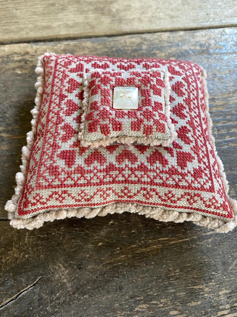 May include: A small, square pincushion with a red and beige cross-stitch pattern. The design features hearts and geometric shapes. A smaller, matching cushion is attached on top, secured with a square button. The edges are trimmed with a beige fringe.