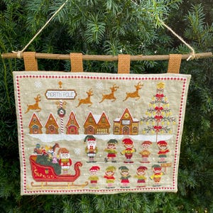 Può includere: Un arazzo natalizio a punto croce con una scena del Polo Nord. Il disegno comprende renne, case, un albero di Natale, Babbo Natale, elfi e una slitta. L'opera è esposta su un tessuto beige, sospesa da un'asta di legno e linguette di tessuto beige.
