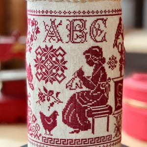 Peut inclure: Un coussin à épingles cylindrique avec un dessus à motifs floraux rouges et blancs. Les côtés présentent des broderies au point de croix en fil rouge sur fond crème, dont les lettres ABC, une femme cousant et un poulet.