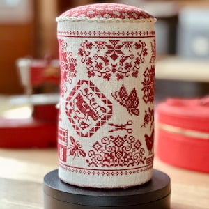 Pu&ograve; includere: Cuscino cilindrico con un disegno ricamato rosso e bianco. La parte superiore presenta un motivo floreale rosso. I lati presentano vari motivi a punto croce rossi, tra cui una casa, una farfalla e motivi floreali. Il cuscino &egrave; su una base nera.
