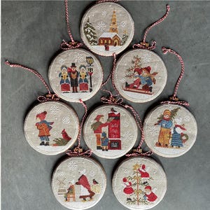 Op de afbeelding: Acht kerstbalornamenten met geborduurde wintertaferelen. Elk cirkelvormig ornament heeft een ander vakantieontwerp, waaronder een kerk, kerstliederen en kinderen die sleeën. De ornamenten zijn versierd met rood en wit touw om op te hangen.