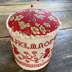 May include: A red and beige pin cushion with three red-topped pins. The top is red with a floral pattern, and the sides are beige with red embroidered lettering and floral designs. A charming sewing accessory.