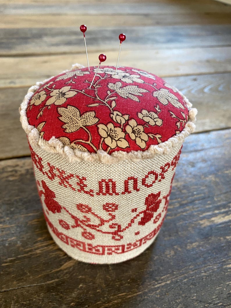 May include: A red and beige pin cushion with three red-topped pins. The top is red with a floral pattern, and the sides are beige with red embroidered lettering and floral designs. A charming sewing accessory.