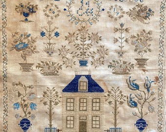 Op de afbeelding: Een vintage borduurwerk met een gedetailleerd geborduurd huis met een blauw dak, omringd door bomen, bloemen en decoratieve elementen. Het ontwerp bevat blauwe en bruine draad op een beige achtergrond, met de initialen "O D" en "J A R".