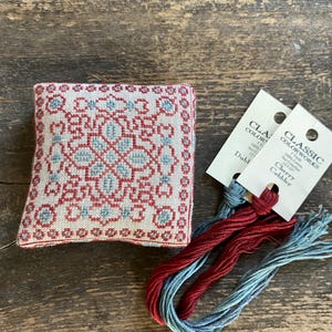 May include: A small, square, embroidered pincushion with a red and blue geometric design. Two skeins of red and blue embroidery floss are next to the pincushion. The floss has tags that read "Classic Colorworks".