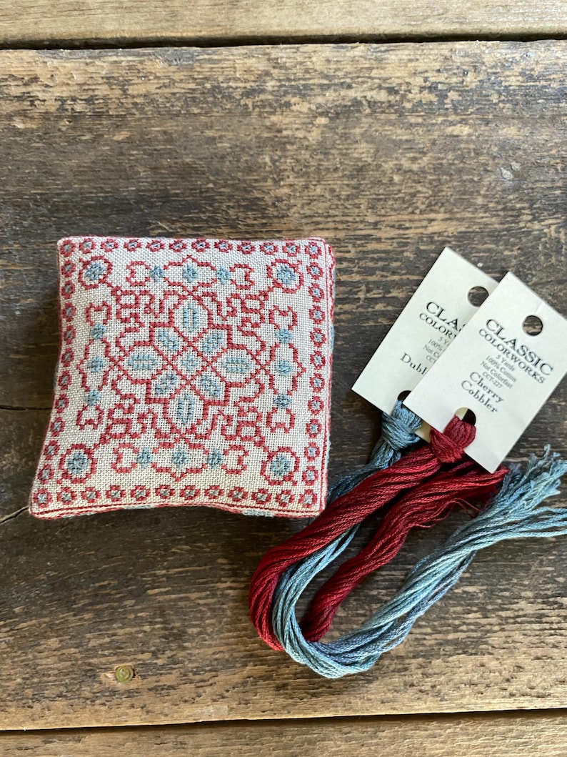 May include: A small, square, embroidered pincushion with a red and blue geometric design. Two skeins of red and blue embroidery floss are next to the pincushion. The floss has tags that read "Classic Colorworks".