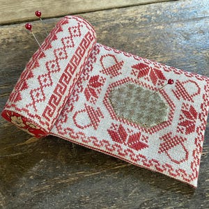 May include: A rolled-up, rectangular pin cushion with a red and cream cross-stitch design. The design includes geometric patterns, floral motifs, and a central patch. Two red-topped pins are inserted into the cushion, which is resting on a wooden surface.