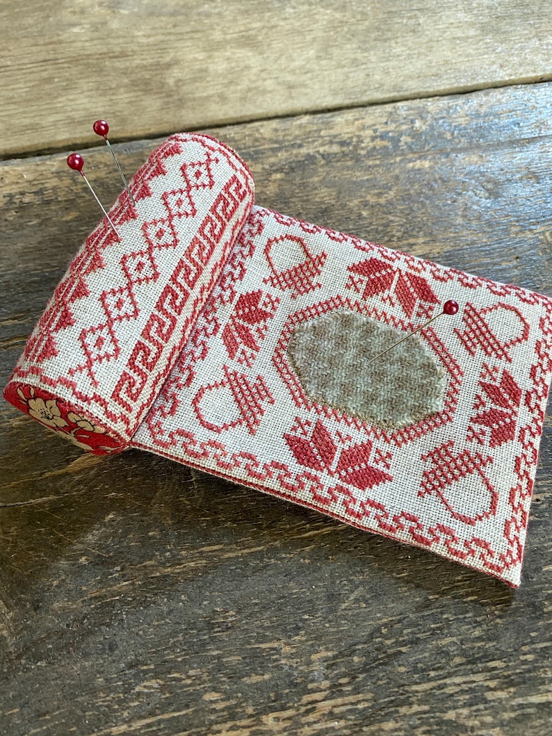 May include: A rolled-up, rectangular pin cushion with a red and cream cross-stitch design. The design includes geometric patterns, floral motifs, and a central patch. Two red-topped pins are inserted into the cushion, which is resting on a wooden surface.