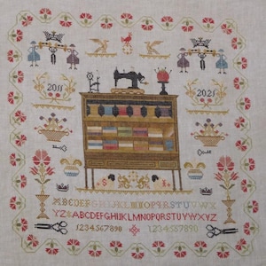 May include: A detailed cross-stitch sampler featuring a sewing machine, a cabinet filled with spools of thread, and decorative floral and bird motifs. The design includes the years 2011 and 2021, along with the alphabet and numbers.