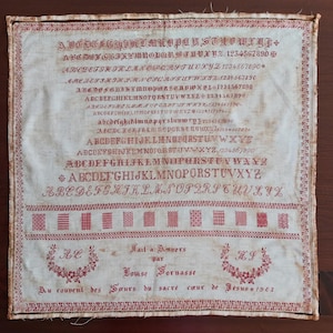 May include: Antique embroidered sampler on cream-colored fabric. The sampler features red embroidered alphabets, numbers, and decorative patterns. Text includes the name Louise Fornasse and the year 1903, with floral accents.