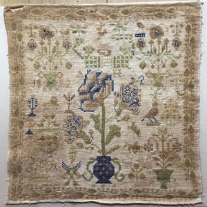 May include: A vintage cross-stitch sampler featuring a central floral design in blue, green, and tan hues. The sampler includes a vase, bird, and various floral and geometric patterns, all stitched on a beige fabric.