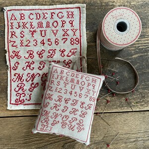 May include: A vintage-style embroidery sampler and a small pillow with red stitched letters and numbers. A spool of dotted ribbon and several pins with red heads are also present, suggesting a crafting theme.