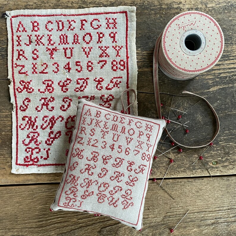 May include: A vintage-style embroidery sampler and a small pillow with red stitched letters and numbers. A spool of dotted ribbon and several pins with red heads are also present, suggesting a crafting theme.
