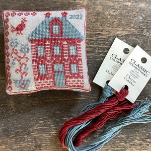 May include: A cross-stitched house ornament with red and blue thread, featuring a house design with the year 2022. Accompanying the ornament are skeins of red and blue embroidery floss with tags that read "Classic Colorworks".
