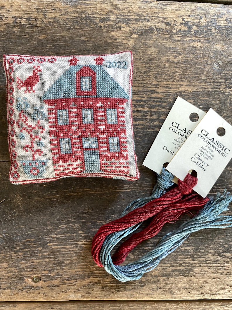 May include: A cross-stitched house ornament with red and blue thread, featuring a house design with the year 2022. Accompanying the ornament are skeins of red and blue embroidery floss with tags that read "Classic Colorworks".