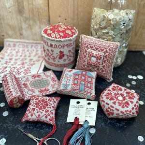 May include: A collection of handcrafted cross-stitch items in red and white, including pincushions, small pillows, and a fabric sampler. A jar filled with buttons and embroidery floss are also present. A pair of scissors is also visible.