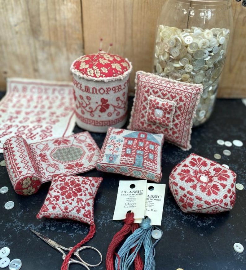 May include: A collection of handcrafted cross-stitch items in red and white, including pincushions, small pillows, and a fabric sampler. A jar filled with buttons and embroidery floss are also present. A pair of scissors is also visible.